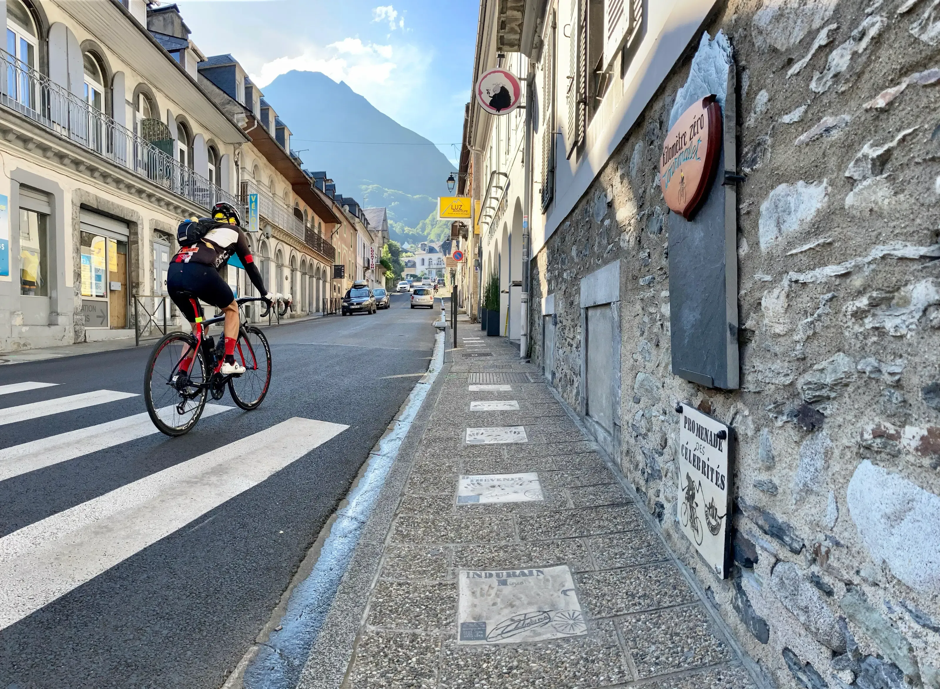 Un cycliste roule dans une rue de village longeant un trottoir doté de plaques commémoratives au sol, avec une haute montagne en arrière-plan.